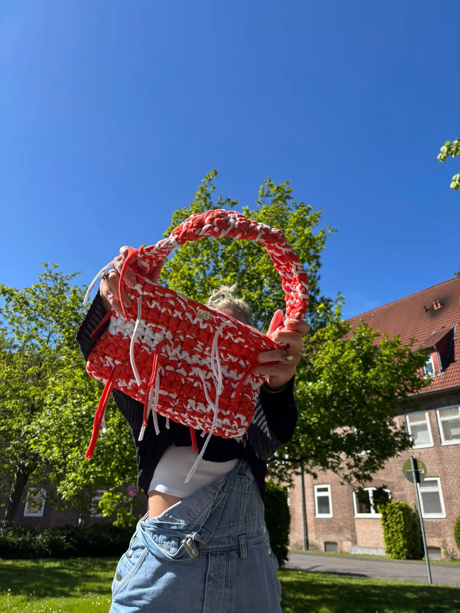 Gehäkelte Tasche in Koralle und Weiß mit Fransen, vor blauem Himmel hochgehalten