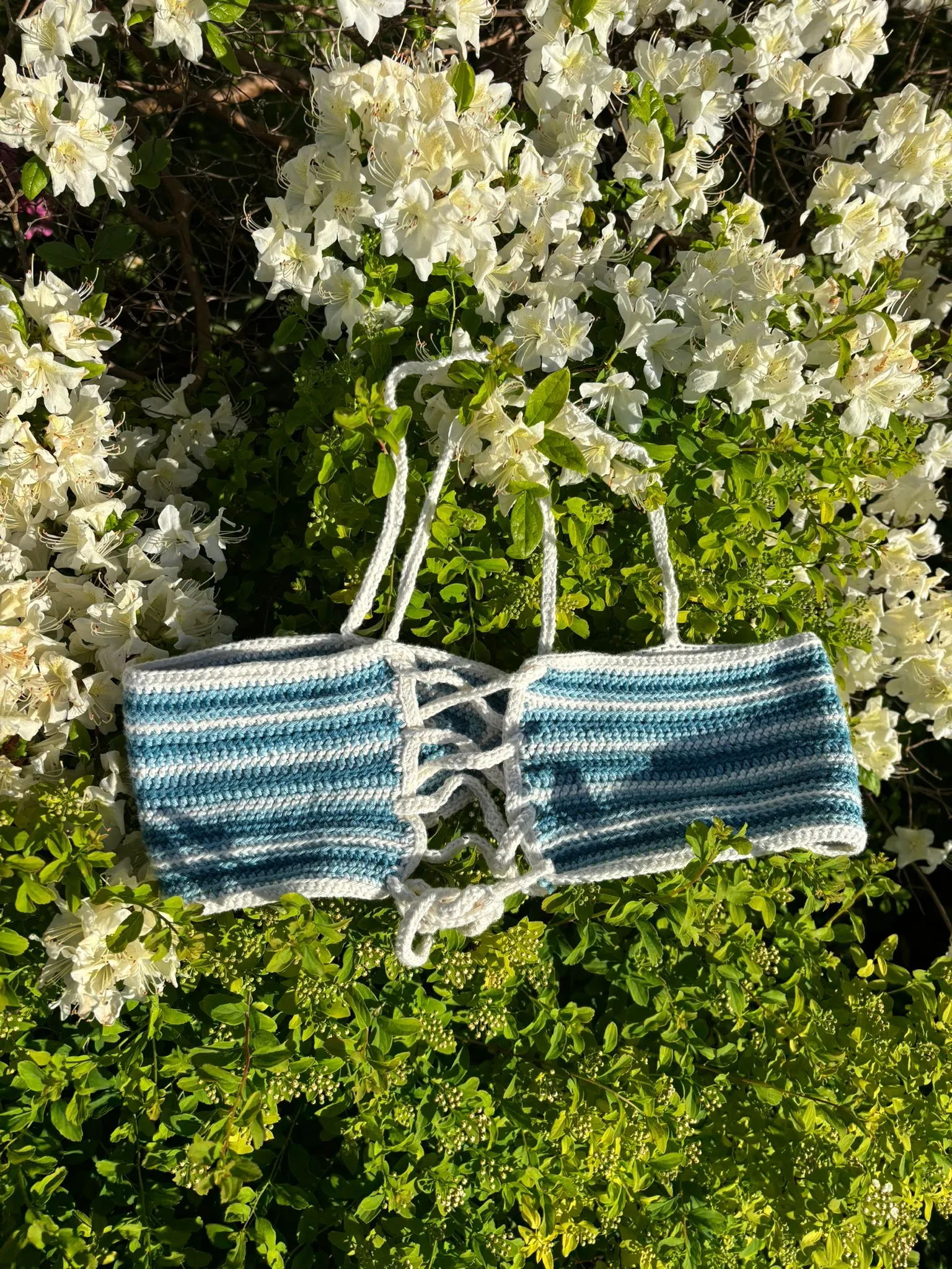 Gehäkeltes Neckholder-Top in Blau und Weiß gestreift, auf blühendem Busch liegend